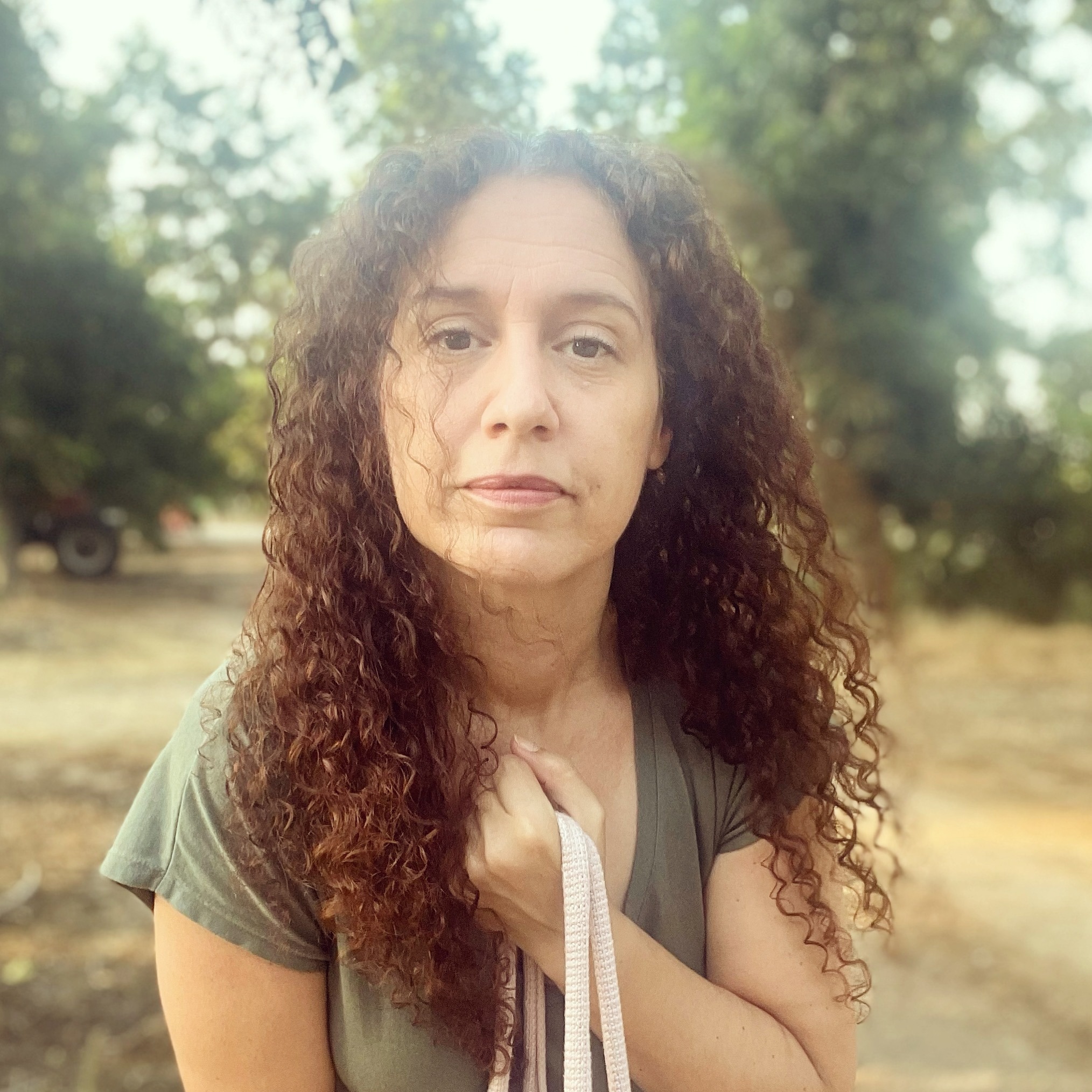 אישה עם שיער מתולתל עומדת בחוץ מול עצים, מחזיקה רצועת שק. מתבוננת ישר למצלמה.